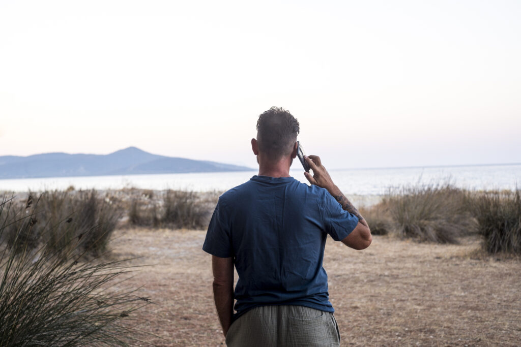 Back view of standing man using mobile phone making phone call in outdoor against a travel scenic destination background with ocean and island. People tourist and cellphone technology connection wifi
